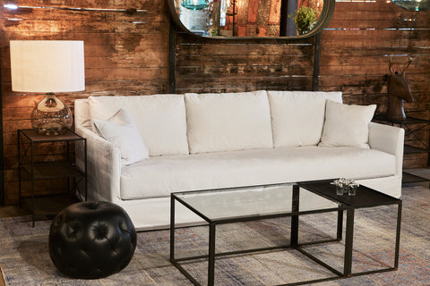 The light colored slipcovered sofa is in a room with dimmed lighting, in front of a dark wood wall with an oval mirror hanging. The Jug Table lamp on the left is on a metal side table. There is a dark leather Pouf Ottoman and a glass and metal Welders coffee table in front. Photographed in Naomi White.