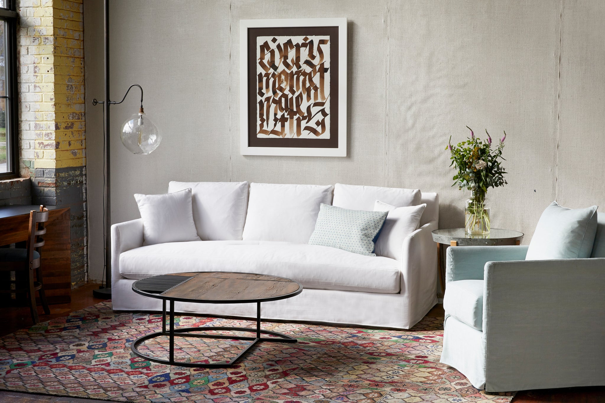  Daytime in an industrial loft. The white slipcovered sofa is in front of a white wall, with a blue slipcovered chair on the right. Floor lamp with a clear glass in the corner. Round coffee table on a multi colored rug. Photographed in Molino White. 