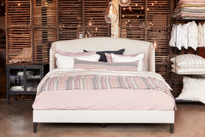  The Emma Bed is shown upholstered with many pillows in white, pink, grey and striped. There is a striped trow on the bed. The legs are in wood with a dark stain. On the left there is a Isaac Nightstand, made of metal with glass doors. On the right there is a shelf with decorative pillows. The background is made with vintage wood crates. Photographed in Avery Oatmeal. 