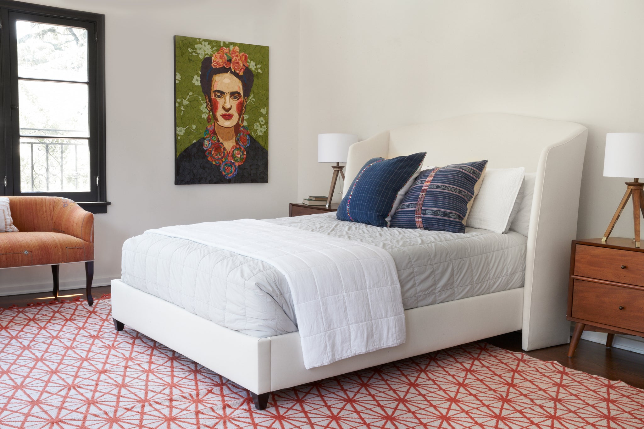  Emma bed in Otis White next to a wood side table. Underneath is a red and white patterned rug. In the background is a white wall room with a painting of a woman. Photographed in Otis White. 