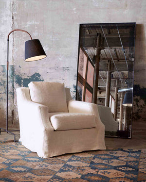  Slip covered chair in white fabric placed next to a floor lamp with dark shade and a floor mirror.  