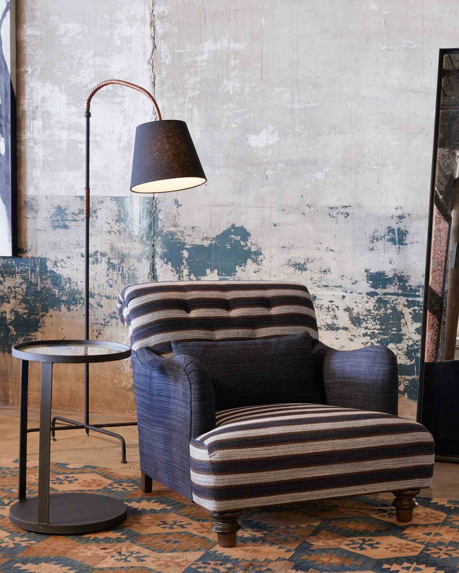  Upholstered chair in blue and white striped fabric, there is a metal side table beside the chair and a floor lamp curved over the chair.  