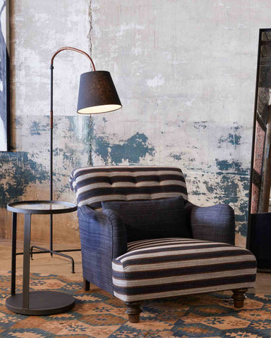 Upholstered chair in blue and white striped fabric, there is a metal side table beside the chair and a floor lamp curved over the chair. 