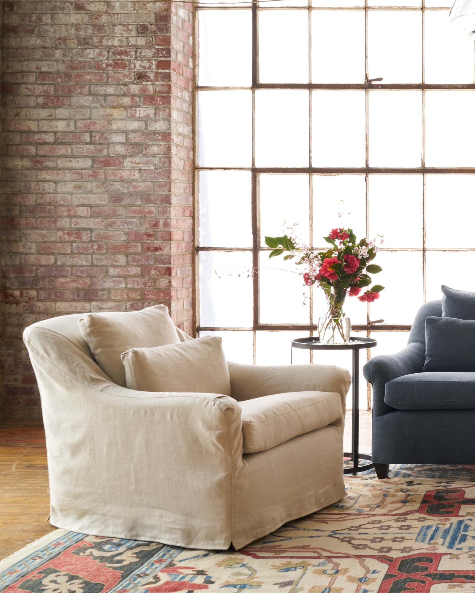  Chair in front of window and brick wall. Blue sofa on the right side and round side table with flowers. Photographed in Noah Bone. 