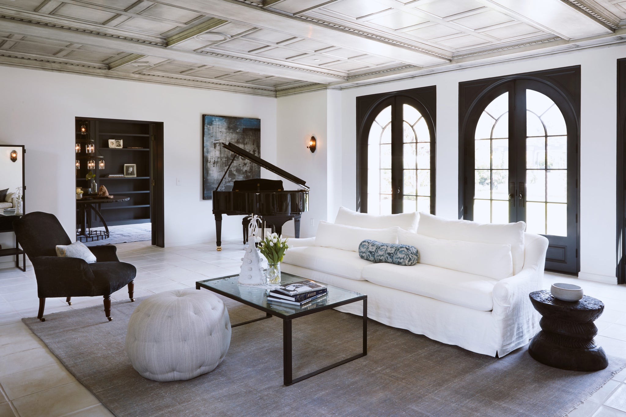  Large living room with daylight with a slipcovered sofa in Brevard Ivory. There is a blackpiano in the back corner, a black armchair on the left side of the room. The coffee table is metal and mirror top, there is a grey ottoman in front. Photographed in Brevard Ivory. 