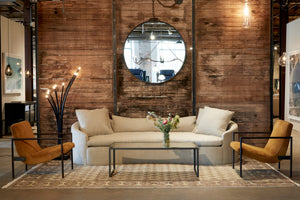  Curved upholstered sofa in Bellamy Oatmeal in front of a wood wall with a round mirror hanging and a Ramo floor lamp on the left. There are two caramel color suede Griffin chairs on each side.The coffee table is metal with a mirror top. Photographed in Bellamy Oatmeal. 