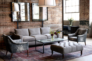  The Gunner sofa is in Rye Warm Grey in a showroom with a wood wall in the back, 2 square mirrors hanging. There are 2 Piper chairs in a grey and white fabric on the sides, a metal coffee table with a glass top in the middle and a rectangular tufted ottoman in grey linen in front. Photographed in Rye Warm Grey. 