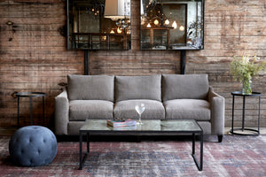  The Gunner sofa is in Rye Warm Grey, in front of a wood wall with 2 square mirrors hanging. Two round side tables on the sides, a rectangular coffee table in metal with a mirror top with a glass of water and book on top. A blue Pouf ottoman is on the left. Photographed in Rye Warm Grey. 