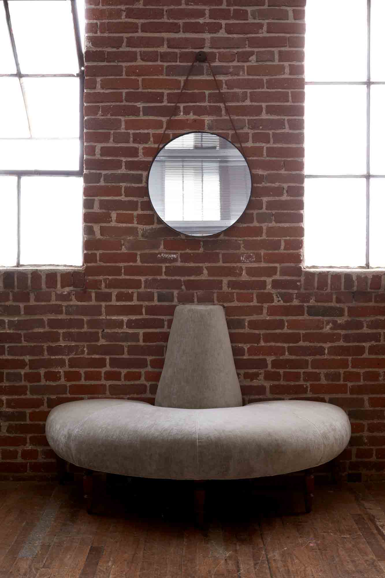  The Half Coin sofa is in Velluto Stone, a natural color velvet, against a brick wall, between 2 windows and a round mirror on the wall. Photographed in Velluto Stone. 