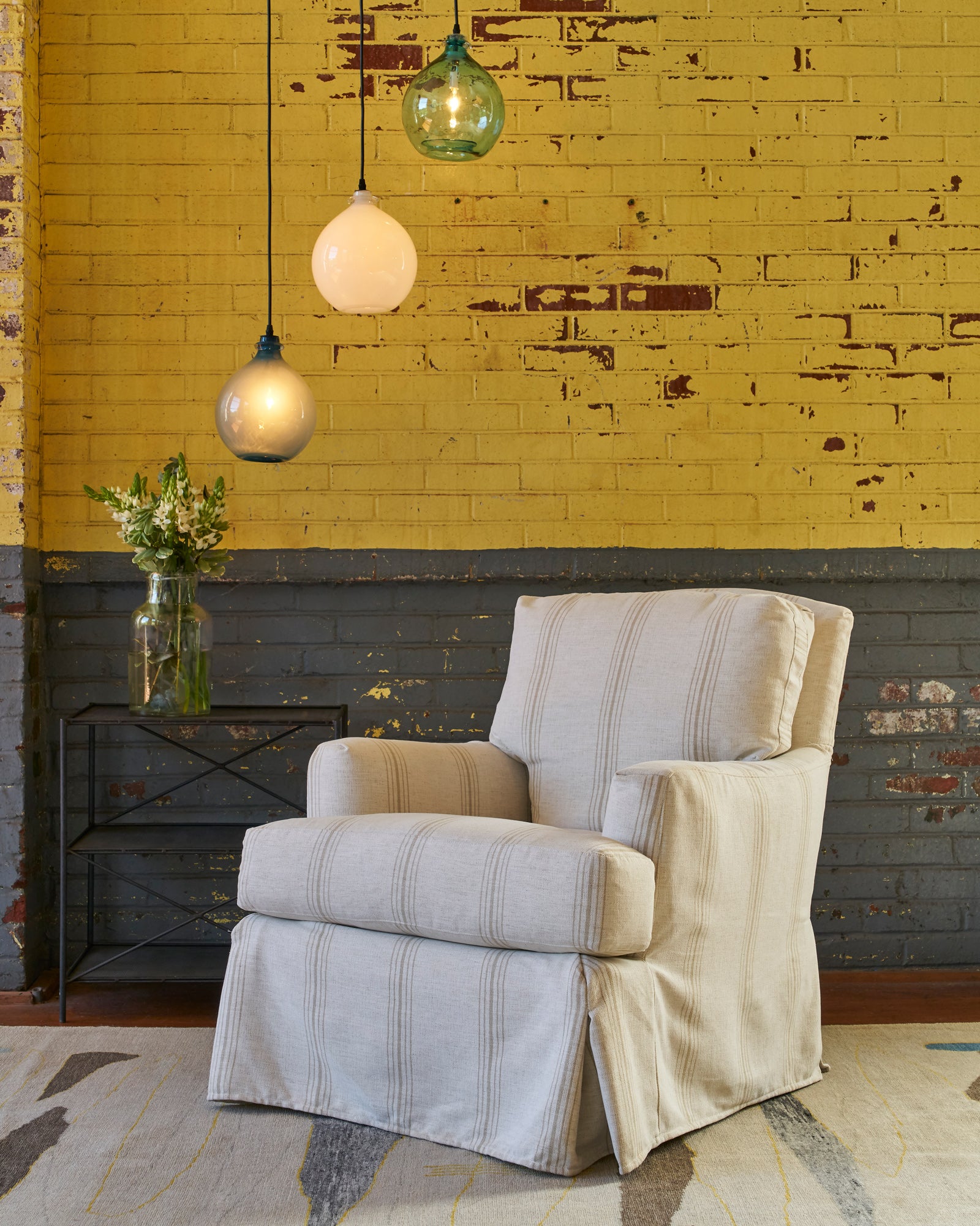  Hampshire Chair slipcovered in Braxton Sand, cream colored fabric with a stripe in front of a yellow and grey brick wall with three pendant lights. Photographed in Braxton Sand. 