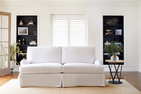 The white slipcovered sofa in Denim White is in a living room with natural daylight. There is a window and two black bookshelves on the back wall. The round wood and metal side table have a vase with green bouquet. Photographed in Denim White