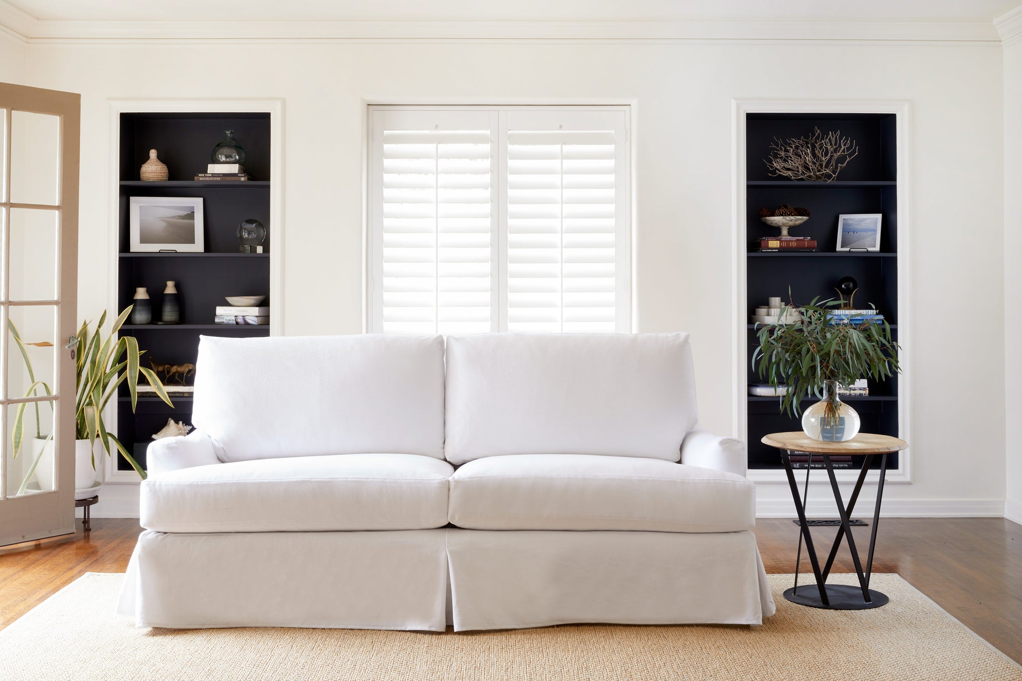  The white slipcovered sofa in Denim White is in a living room with natural daylight. There is a window and two black bookshelves on the back wall. The round wood and metal side table have a vase with green bouquet. Photographed in Denim White. 