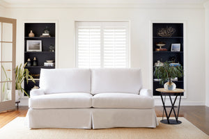  The white slipcovered sofa in Denim White is in a living room with natural daylight. There is a window and two black bookshelves on the back wall. The round wood and metal side table have a vase with green bouquet. Photographed in Denim White. 