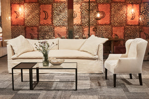  Slipcovered Havana sofa in a natural cream color fabric. Sitting in front on a vintage Tapa Cloth, with showroom lighting. The coffee table is metal and glass with a bowl and flowers on it. The Butterfly chair by John Derian in a light color fabric is on the right. Photographed in Vanocur Natural. 