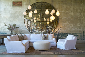  Large white sectional in front of an oversized mirror. A round ottoman in front and a white chair on the right. White glass pendant lights hang from the ceiling. Photographed in Lan Ivory. 