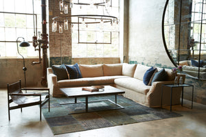 Hayden Deluxe 2 arm Sectional in Quixote Oatmeal sits next to a metal side table, a wood coffee table, and a leather chair. Underneath the sectional is a patterned dark rug. Above the sectional is a chandelier. Photographed in Lan Oatmeal 