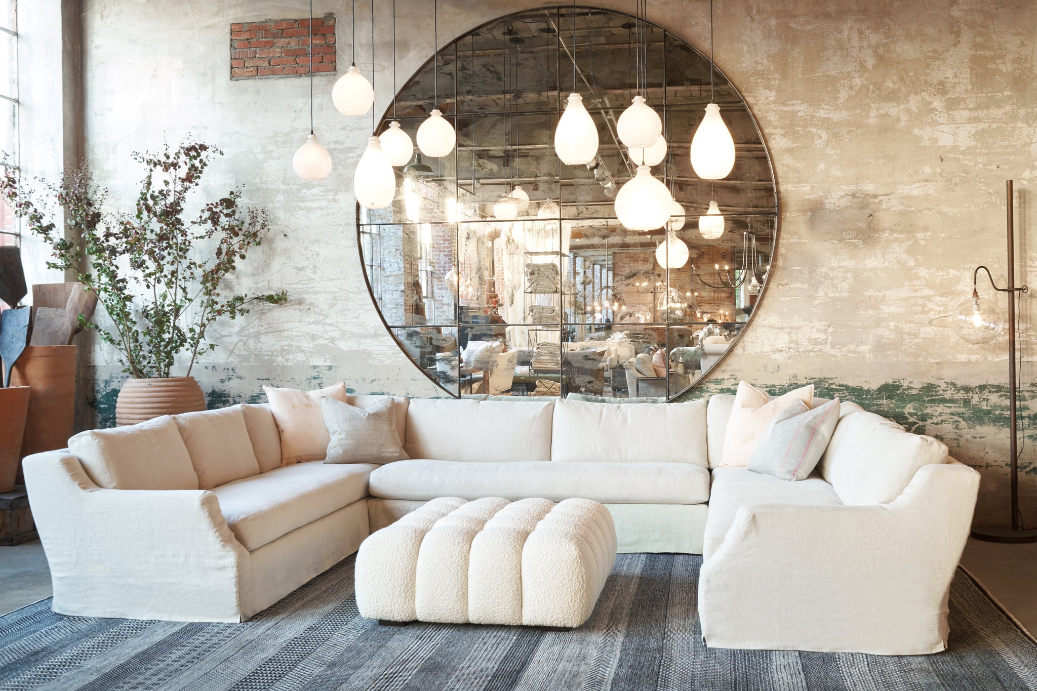  Large 3 piece sectional in front of a round mirror. A cream colored ottoman is in the middle. There are white glass light pendants hanging above. Photographed in Wooly White. 