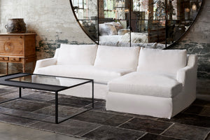  Hayden Deluxe 2pc Sectional in Lan Ivory next to a glass coffee table. Underneath the sectional is a dark rug. In the background is a large mirror that hangs on the wall. Photographed in Brevard Ivory. 