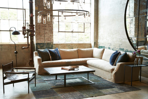 Hayden Deluxe 2 arm Sectional in Quixote Oatmeal sits next to a metal side table, a wood coffee table, and a leather chair. Underneath the sectional is a patterned dark rug. Above the sectional is a chandelier. Photographed in Lan Oatmeal.