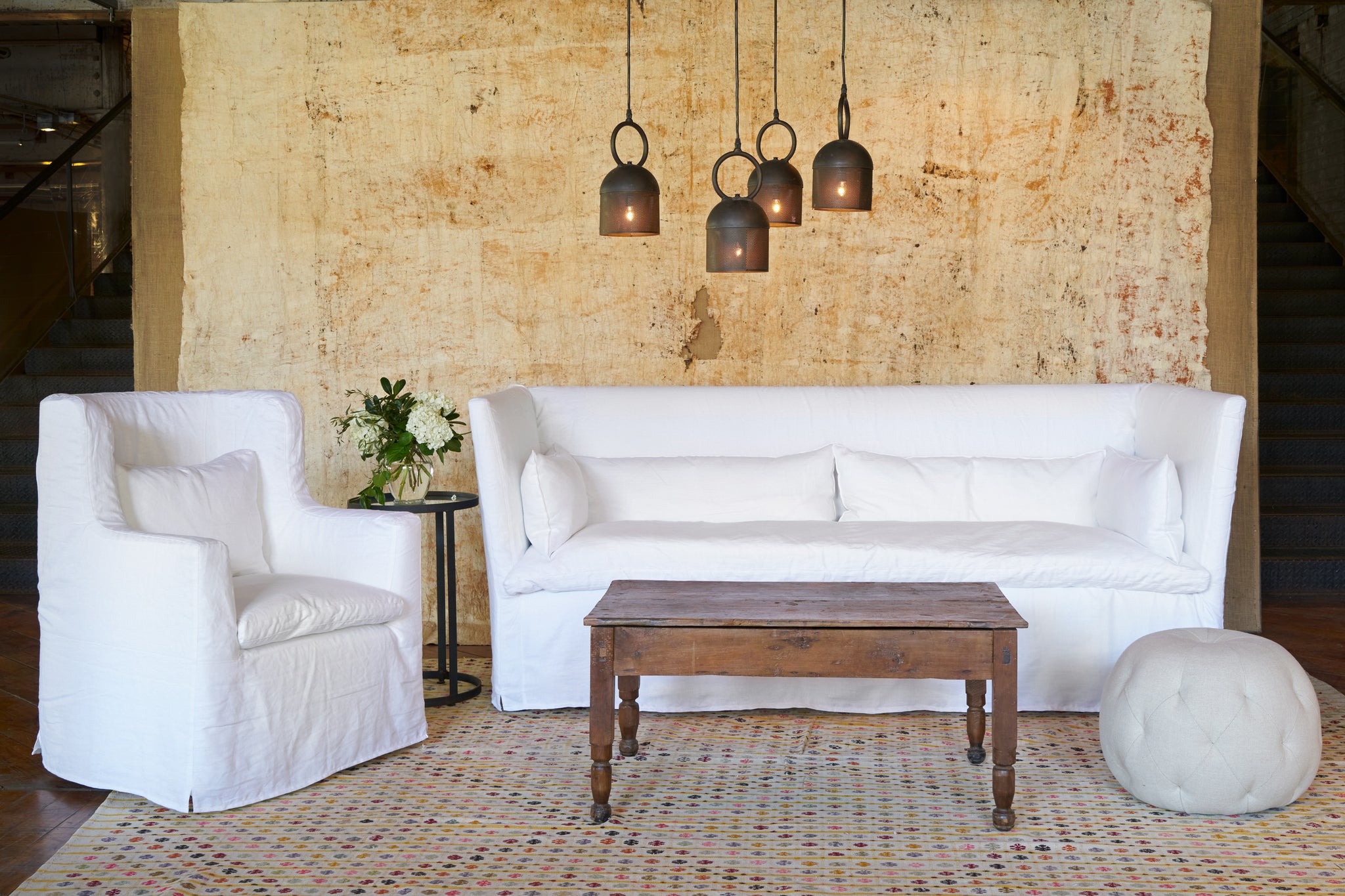  White chair on the left of a white sofa in front of a yellow backdrop and 4 metal light pendants. A wood coffee table and a tufted pouf ottoman are in front. Photographed in Otis White. 
