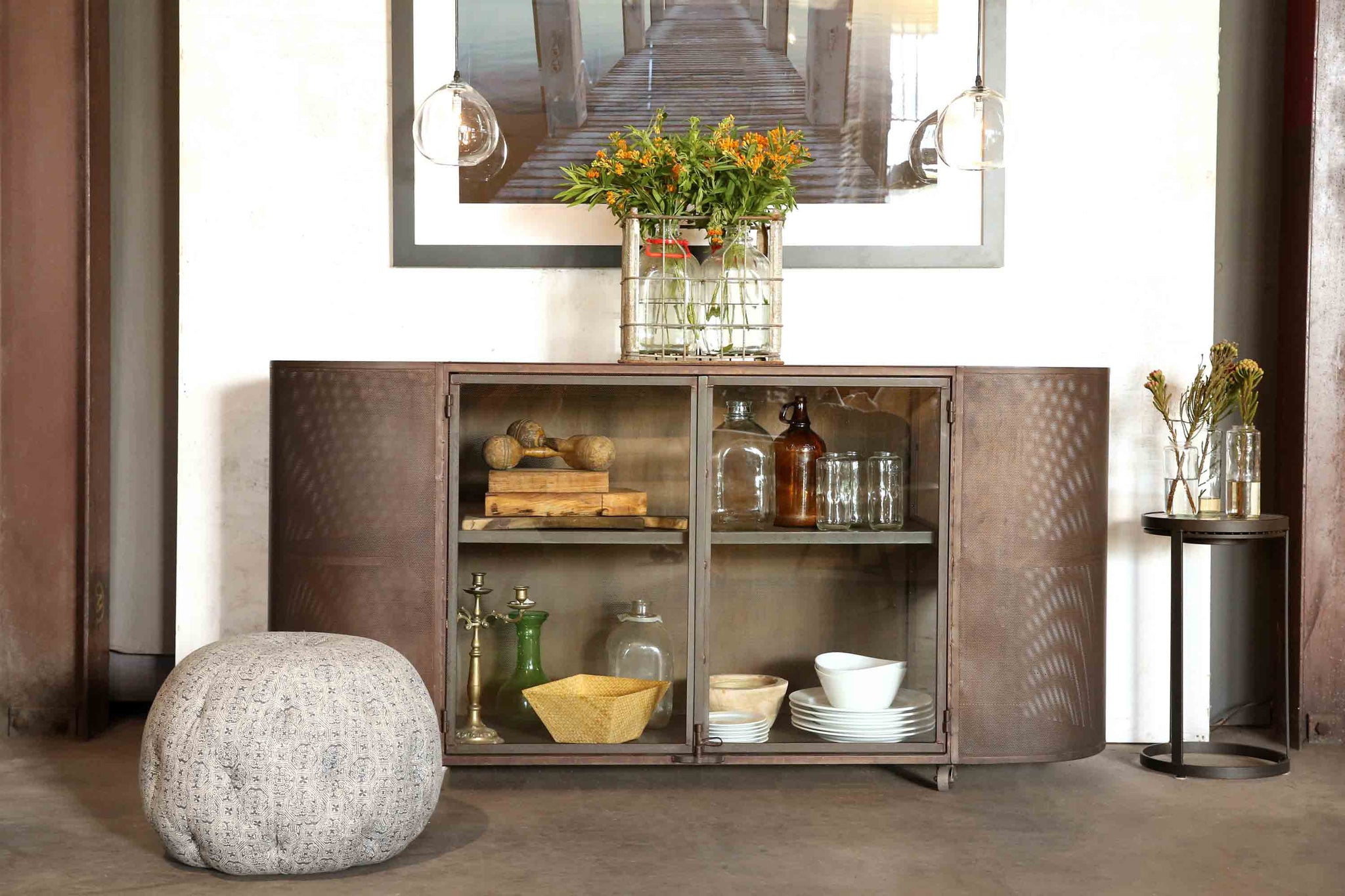  Oval issaac credenza with two teardrop pendants in clear hanging above. Rotor side table beside the issaac credenza and a pouf ottoman in printed fabric. 