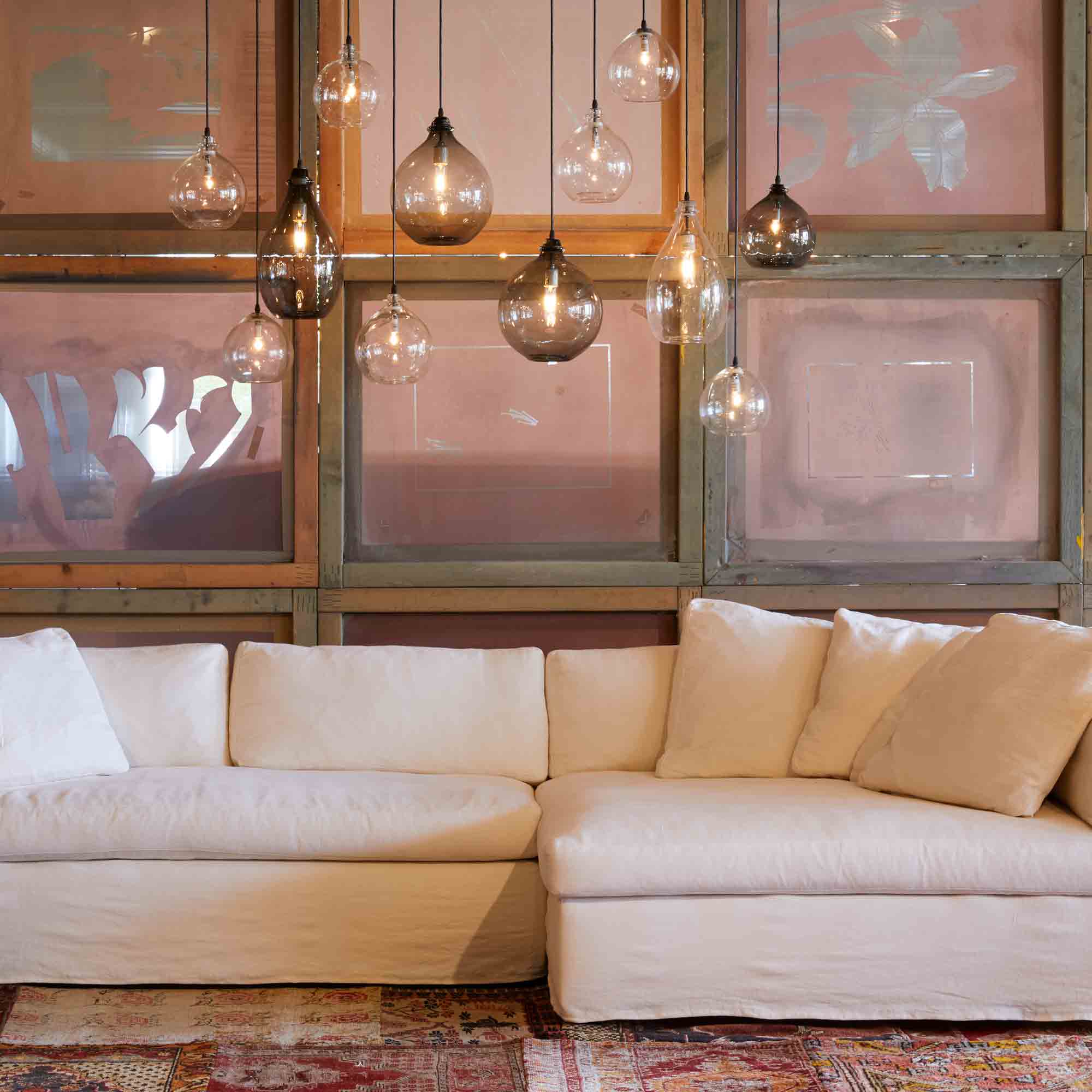  Slipcovered sectional in light neutral fabric with combination of jug pendants hanging above in clear and smoke finish. Repurposed silk screens used as a background wall.  