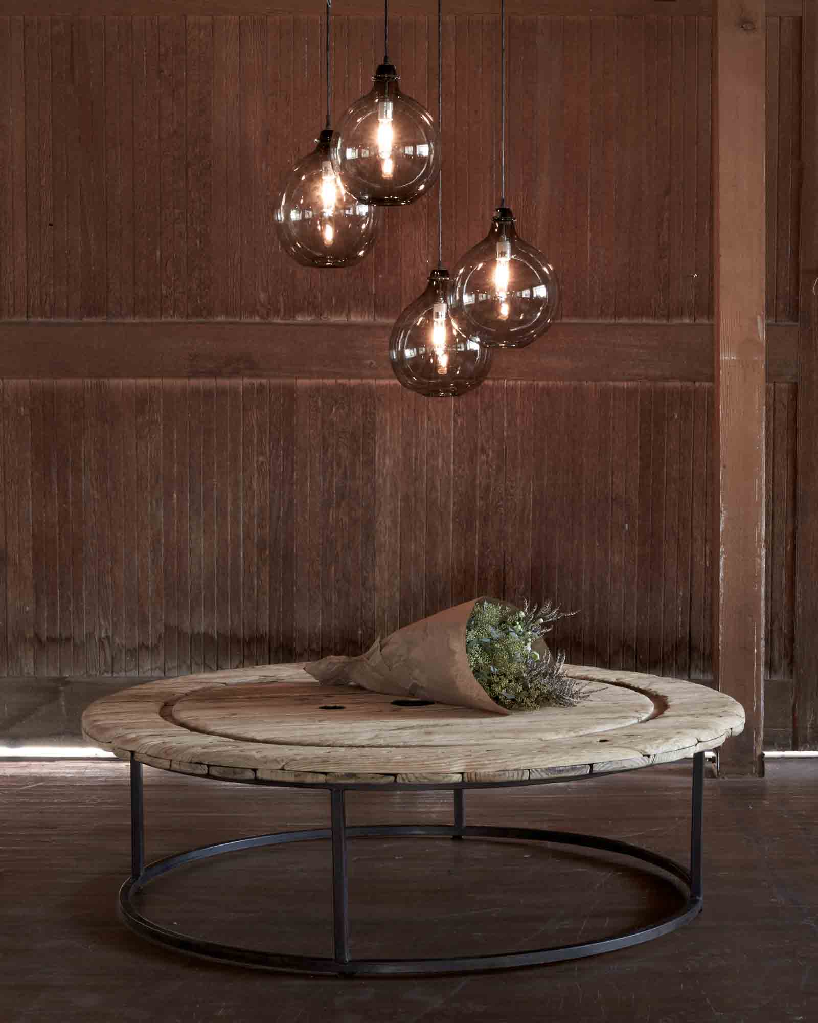  Wood round coffee table with four Jug Lamps size large in smoke finish hanging above coffee table. Wood wall in the background and bouquet of flowers on the coffee table.  