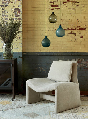 Upholstered chair in neutral fabric and three small jug lamps hanging above in smoke and ice finish. Setting is placed against distressed brick wall painted dark blue and yellow.  