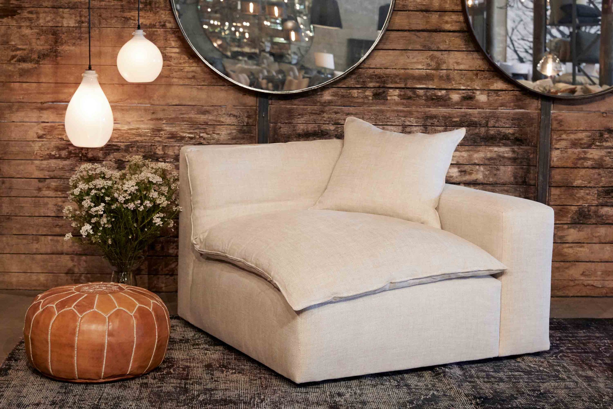  Upholstered corner sofa piece in light neutral fabric with leather ottoman and one jug oval lamp and jug lamp in snow finish. Wood wall with two round wall mirrors.  