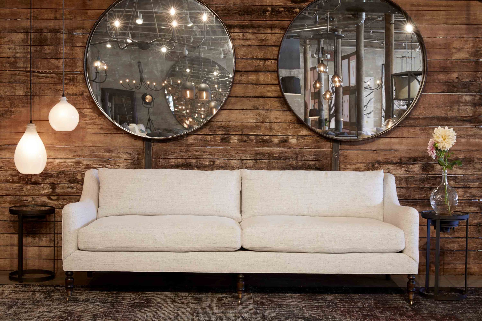  Upholstered sofa in light neutral fabric with two rotor side tables on either side and one jug oval and jug small lamp hanging above one side table in snow finish. Pieces are in front of a wood wall with two large round wall mirrors hanging. 