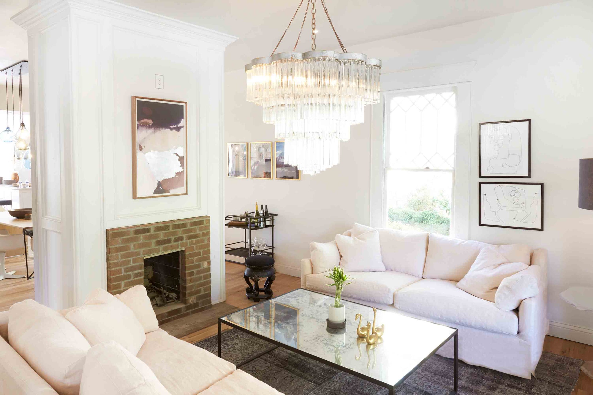  Small living with daylight and two facing Lanister sofas in Brevard Rose linen. A large glass chandelier is hanging above a mirror coffee table in front of a brick fireplace. Photographed in Brevard Rose. 