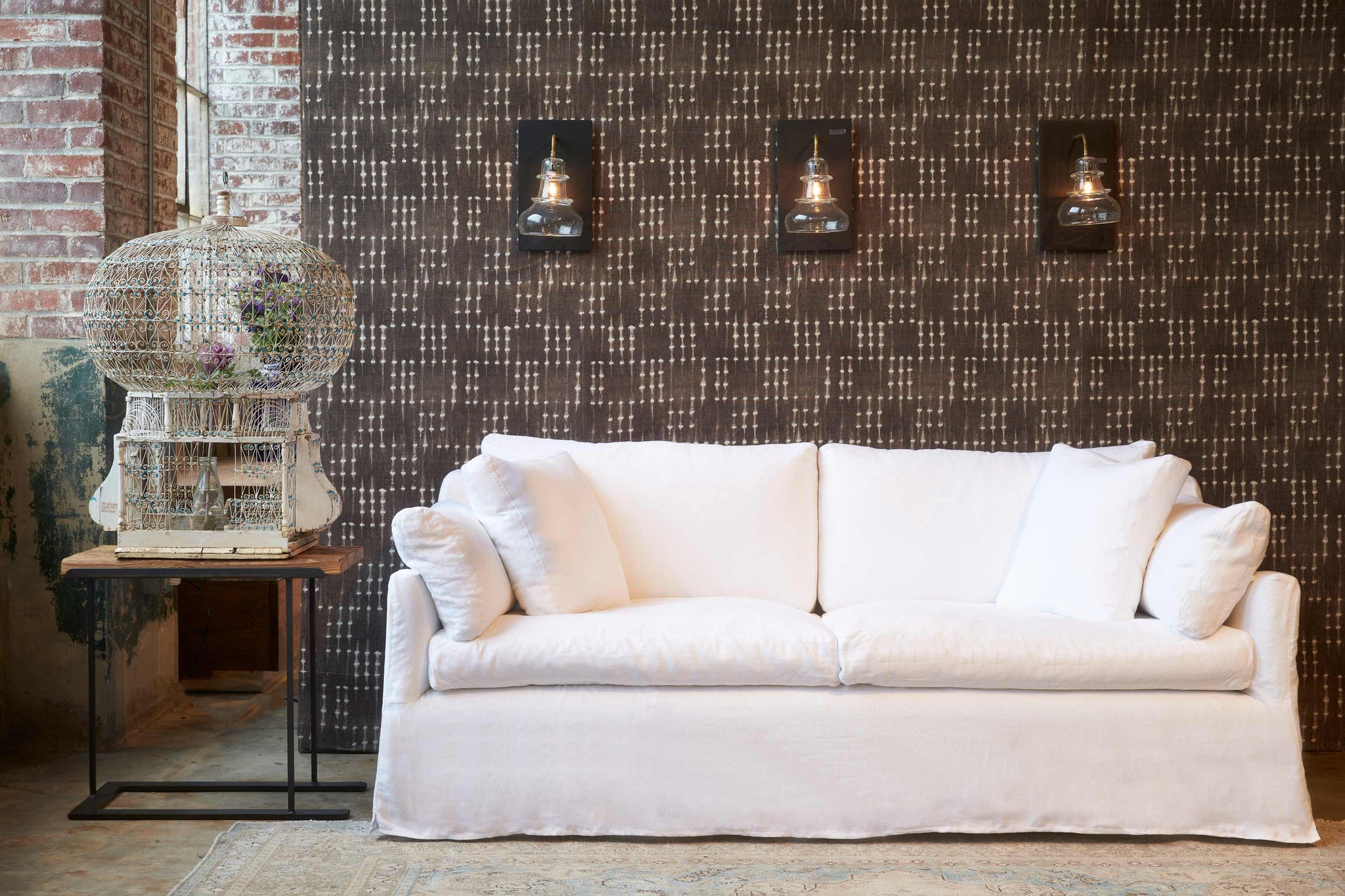  Lanister sofa in Otis White in front of a brown and white fabric wall. Three light sconces on the wall and a large vintage white birdcage is on the left. Photographed in Otis White. 