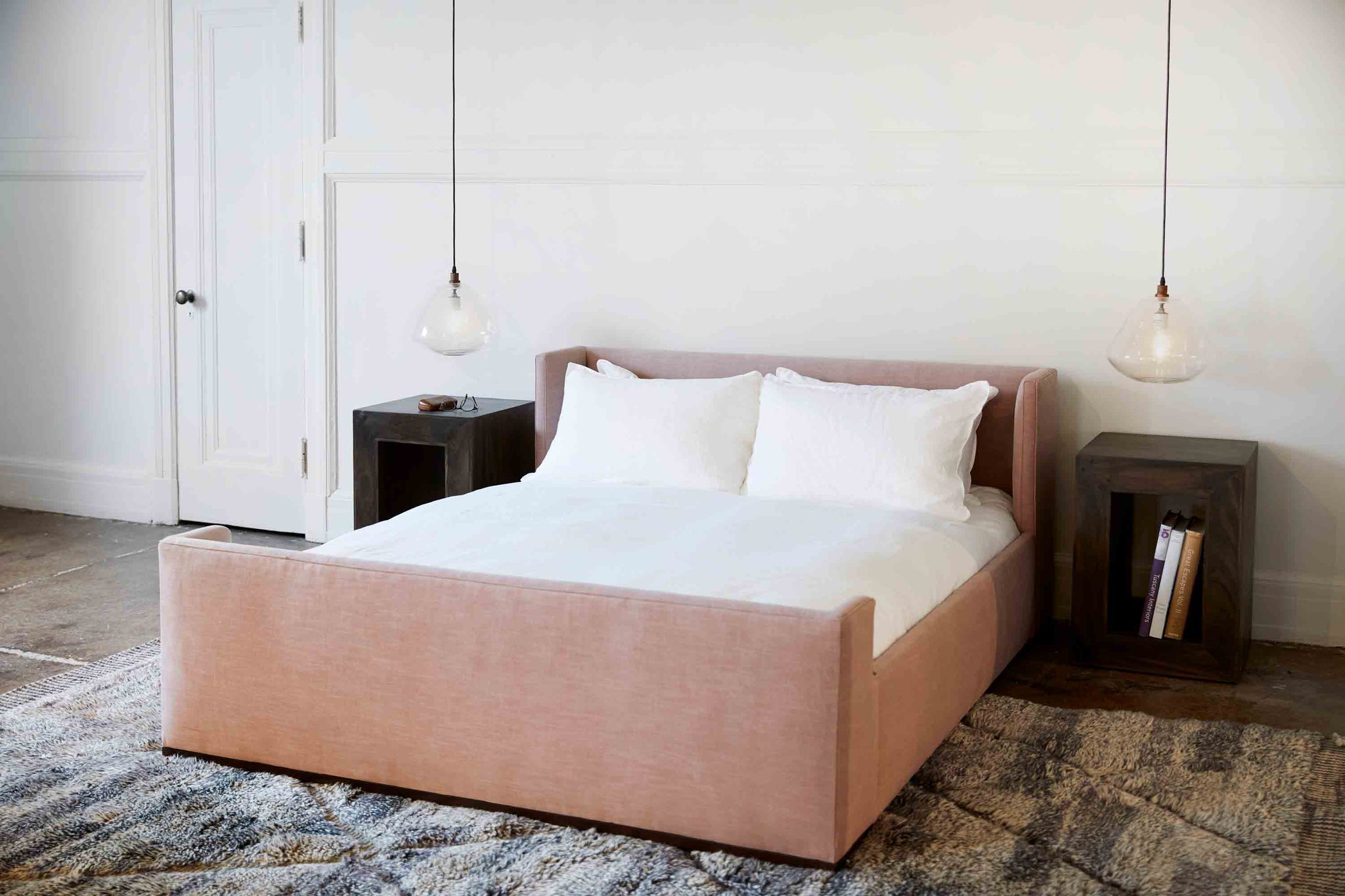  Laurel bed in Molino Blush next to two wood side tables. Underneath is a patterned rug. On top of the side tables are clear lamps that hang above. Photographed in Molino Blush. 