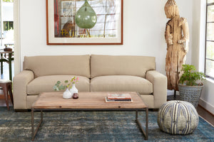  In a living room with daylight, the Loft sofa in Naoki Hemp is in front of a white wall with a painting on the wall and a green light hanging from the ceiling. There is a wood sculpture in the right corner, a plant in a metal bucket and a Pouf ottoman in blue and white fabric on the right of a wood coffee table. Photographed in Naomi Hemp. 