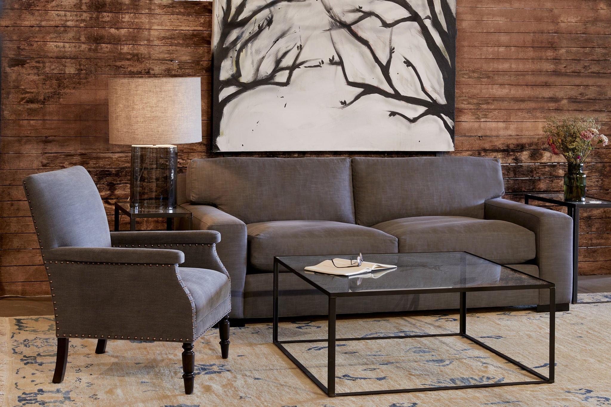  In a showroom with moody lighting, the Loft sofa is in Molino Slate, in front of a wood wall with a black and white painting on the wall. Metal and glass side tables with a Cylinder table lamp on the left and flowers on the right side. A square metal and glass coffee table in front with a book vase and reading glasses on top. The Hailey Chair in Molino Slate is on the left. Photographed in Molino Slate. 