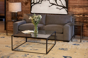  In a showroom with moody lighting, the Loft sofa is in Molino Slate, in front of a wood wall with a black and white painting on the wall. Metal and glass side tables with a Cylinder table lamp on the left. A square metal and glass coffee table in front with a glass vase with white flowers. Photographed in Molino Slate. 