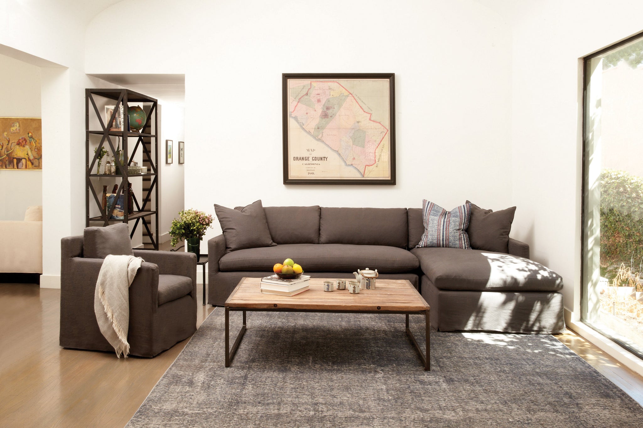  Louis Sectional in Logan Grey next to a chair and wood coffee table. Underneath the sectional is a grey rug. In the background are white walls with a painting hanging from the wall. Photographed in Logan Grey. 