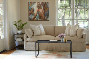  Living room daytime, the Louis sofa is in Brevard Burlap, in front of a large window and a wall with a painting hanging. There is a metal side table on the left, a metal coffee table in front. Two decorative pillows on the sofa. Photographed in Brevard Burlap. 
