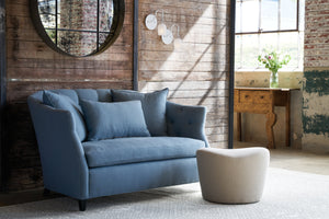  A blue loveseat in front of a wood wall with a white ottoman in front. Two white sconces are on the wall next to a round mirror. In the background, there is a carved wood console with flowers on top. Photographed in Brevard Ciel. 