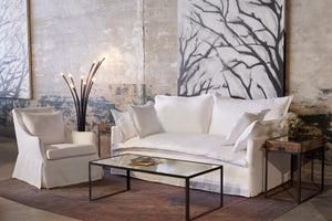  Lucy chair in Otis White next to a light sofa, a metal floor lamp, and glass coffee table. In the background is a concrete wall with a large painting hanging. Photographed in Otis White 