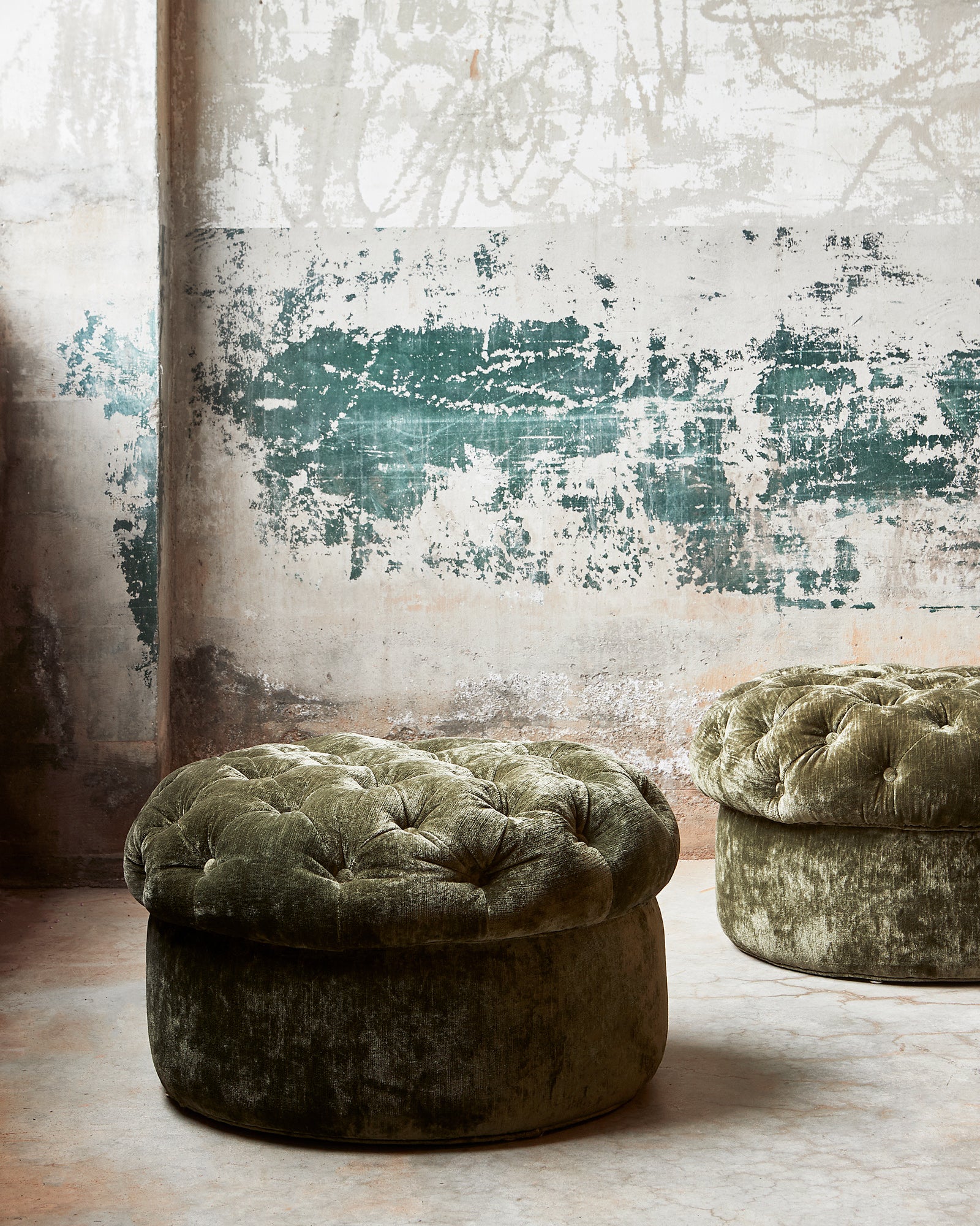  Two green velvet tufted ottomans in front of a patinaed concrete wall. Photographed in Velluto Olive. 