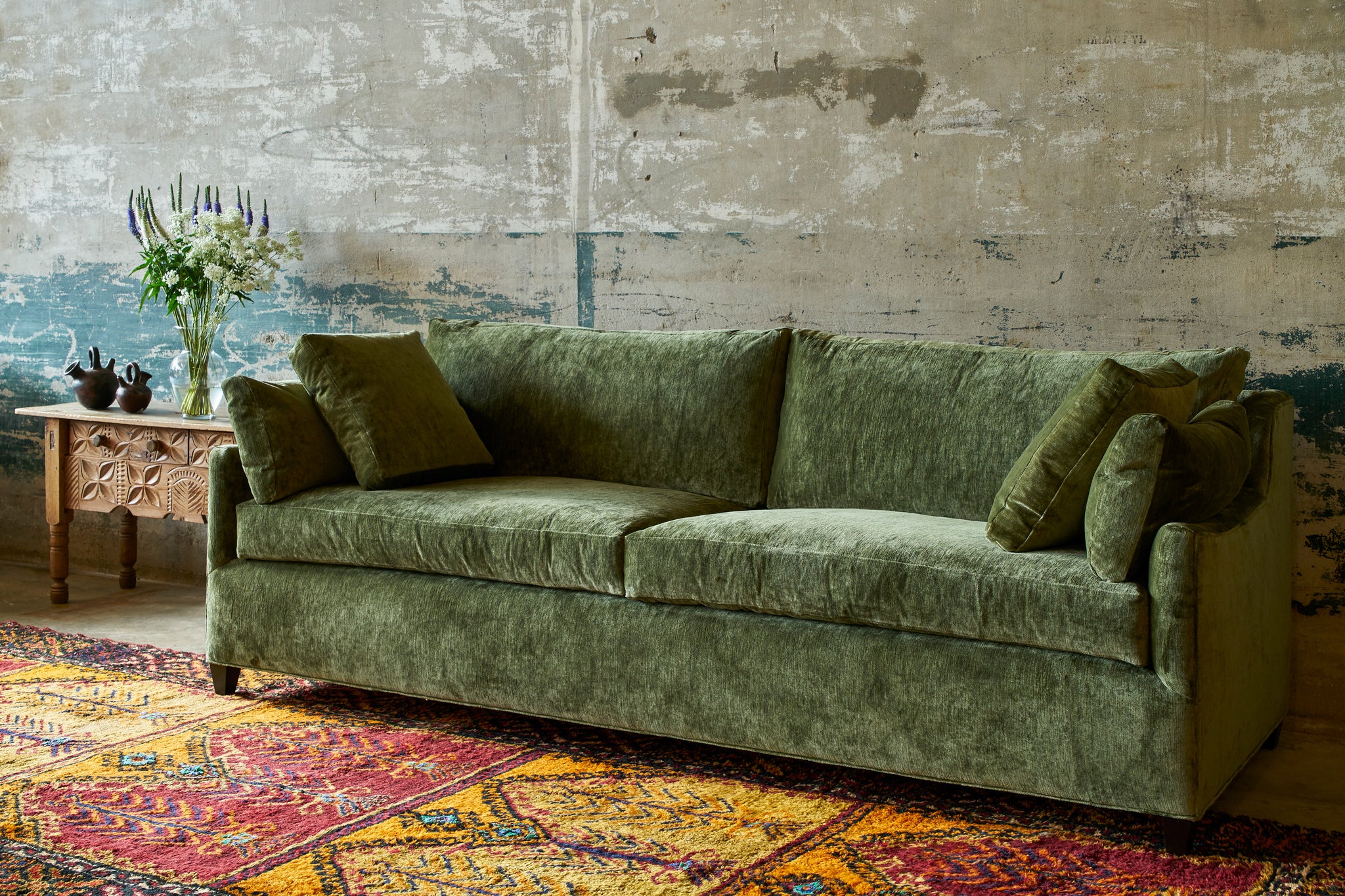  Milo Sofa in a green velvet in front of a concrete wall and a Guatemalan wood side table with flowers on top. Photographed in Velluto Olive 