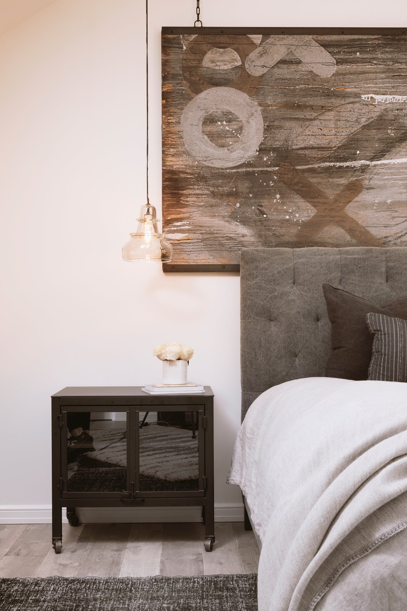  Zoomed in bedroom shot showing corner of upholstered bed in light grey fabric, issaac night stand with morse pendant hanging above in clear color. 