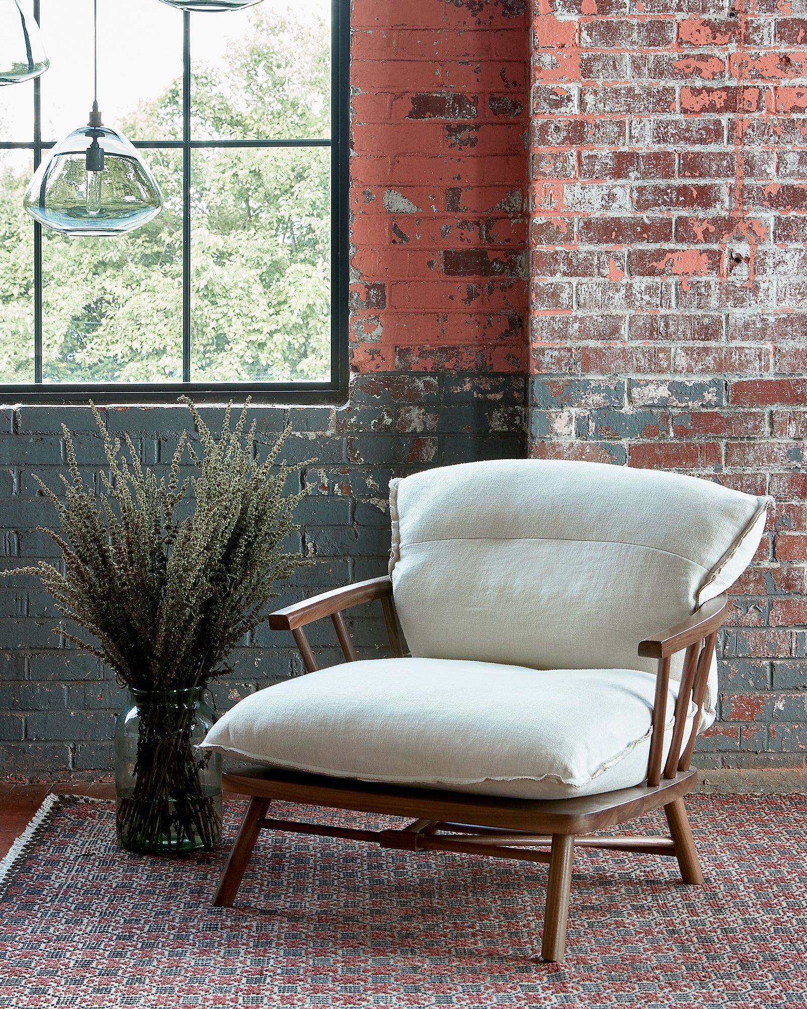  Chair with a wood frame in front of a brick wall with a vase on the floor on the left with plants. Photographed in Brevard Birch. 