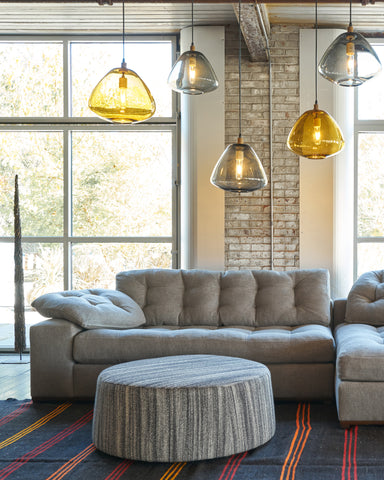 Ollie ottoman on in grey striped fabric in front of a sectional. Glass light pendants are hanging from the ceiling. Photographed in Veneto Black.