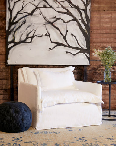 Paloma chair and a half in Otis White next to a dark pouf and metal side table. In the background is a wood wall with a large painting of tree branches. Photographed in Otis White.