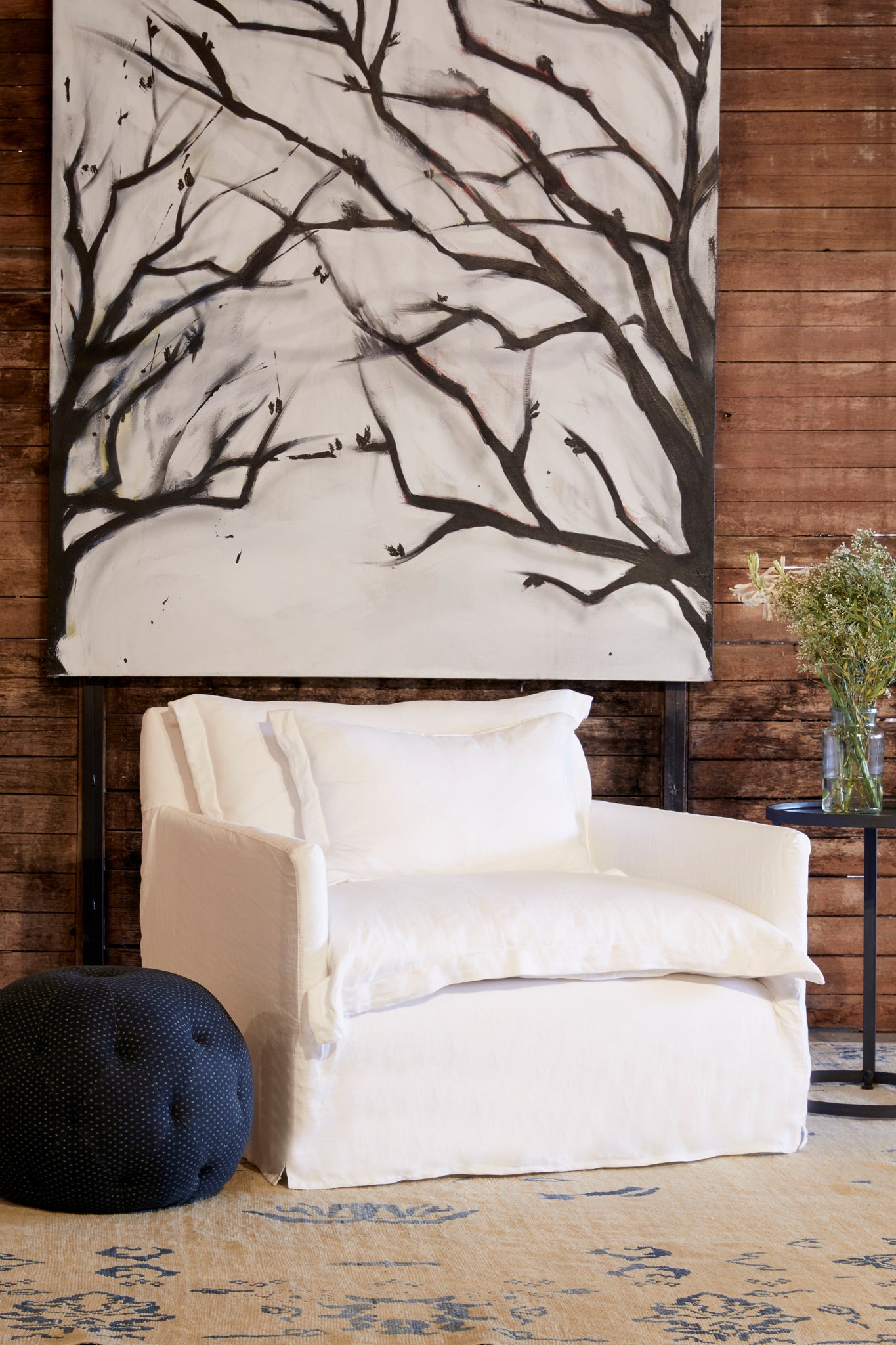  Paloma chair and a half in Otis White next to a dark pouf, and a metal side table. In the background is a wood wall with a large painting of tree branches. Photographed in Otis White. 