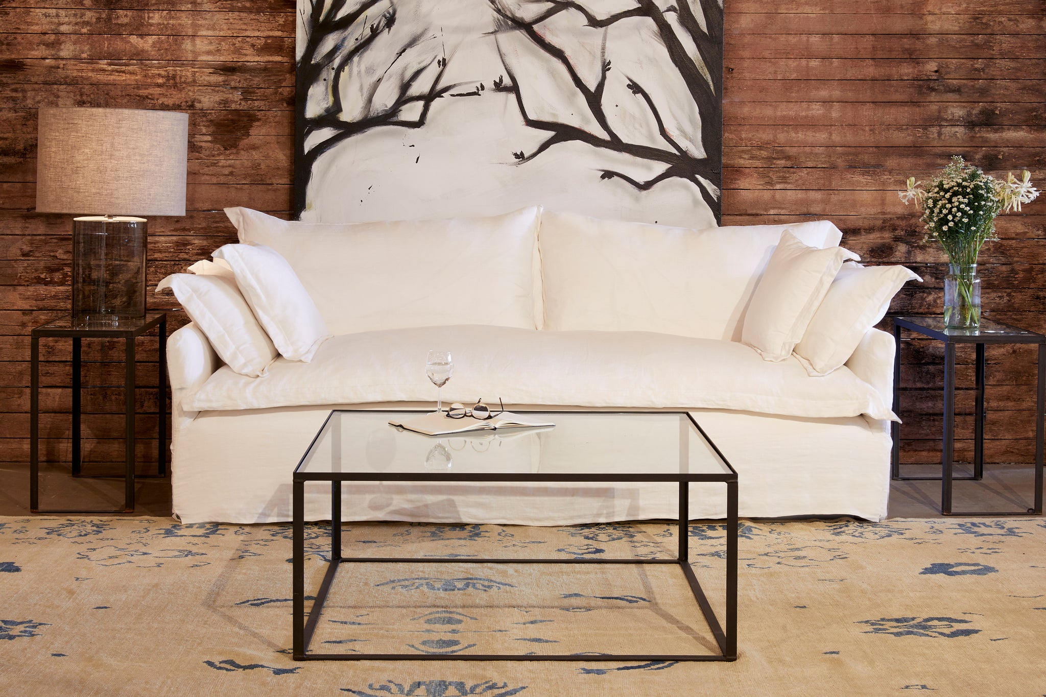  Paloma sofa slipcovered in front of a wood wall with a black and white painting of a tree. The Welders coffee table is in front, with a glass top, The Lena side tables have a Cylinder table lamp on the left and some white flowers on the right. Photographed in Otis White. 