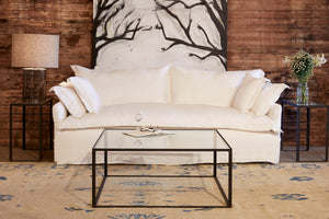  Paloma sofa slipcovered in front of a wood wall with a black and white painting of a tree. The Welders coffee table is in front, with a glass top, The Lena side tables have a Cylinder table lamp on the left and some white flowers on the right. Photographed in Otis White. 