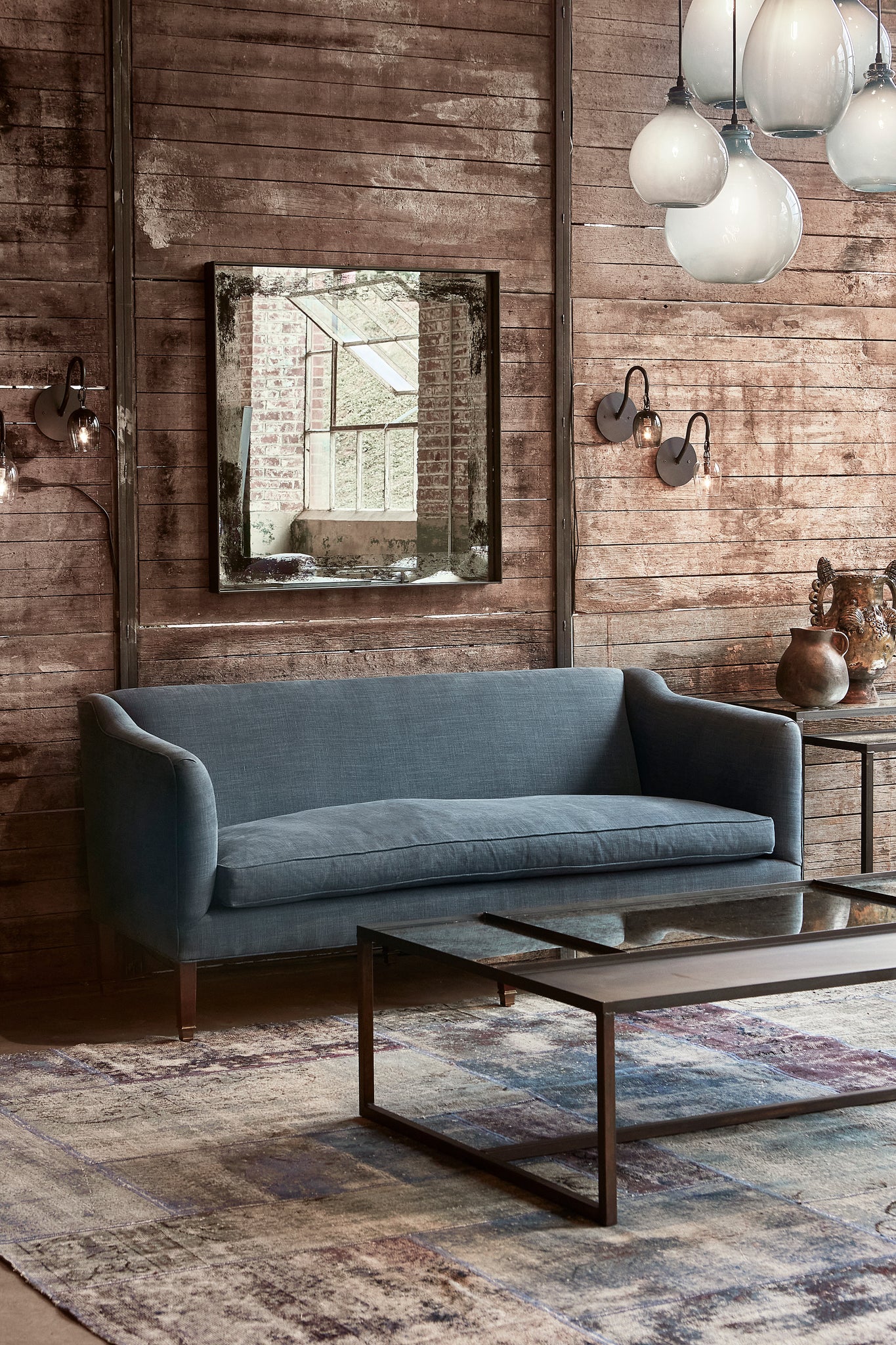  Upholstered sofa in dark grey/blue fabric with rectangular coffee table in front. Setting placed in front of wood distressed wall with square wall mirror and two pear sconces on either side. 